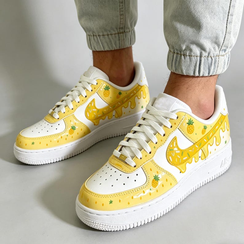 May include: White and yellow sneakers with a pineapple design. The shoes feature a white base with yellow accents, including a dripping pineapple design on the Nike swoosh and sides. Small pineapple and star details are scattered across the shoe.
