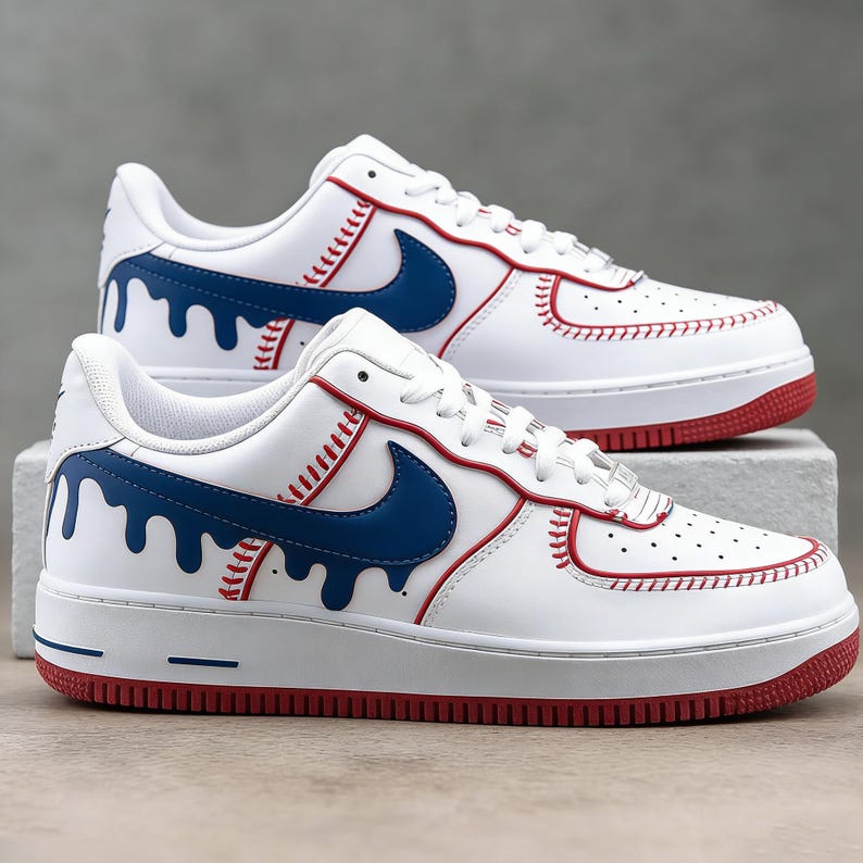 May include: White sneakers with red and blue accents. The shoes feature a white base with a blue swoosh and a red sole. The design incorporates baseball-inspired stitching and a dripping blue paint effect.