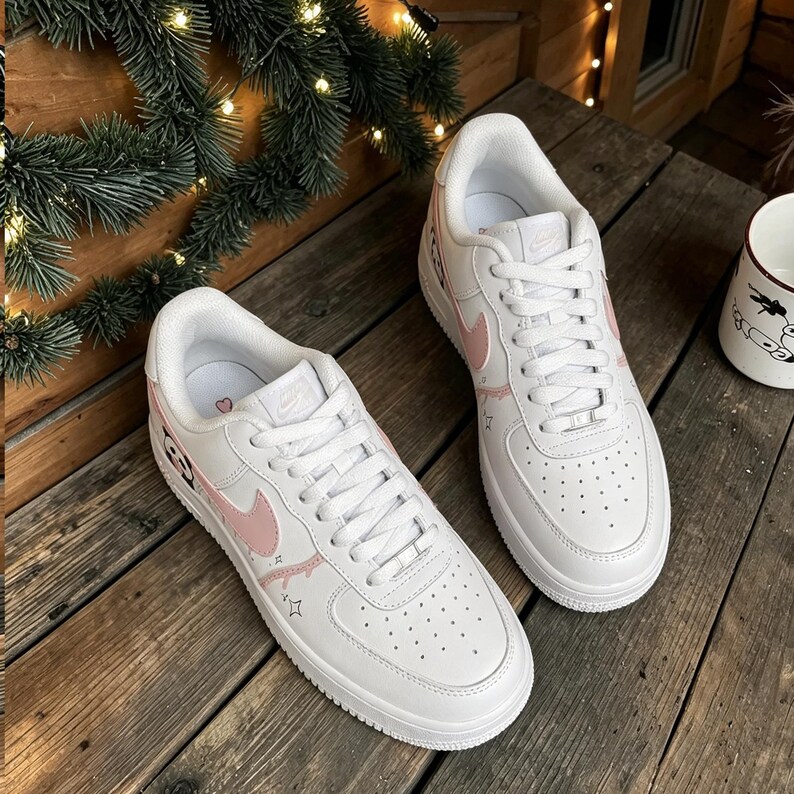 Puede incluir: Un par de zapatillas blancas con detalles en rosa, incluyendo el logo de Nike y elementos decorativos. Las zapatillas est&aacute;n sobre una superficie de madera, mostrando un dise&ntilde;o deportivo cl&aacute;sico con un toque moderno.