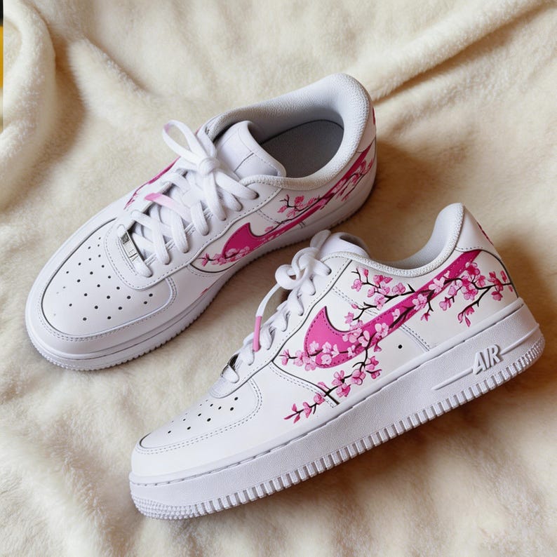 May include: White sneakers with pink cherry blossom design. The shoes feature a pink Nike swoosh and cherry blossom branches across the sides. The soles are white with the word "AIR" on the side.