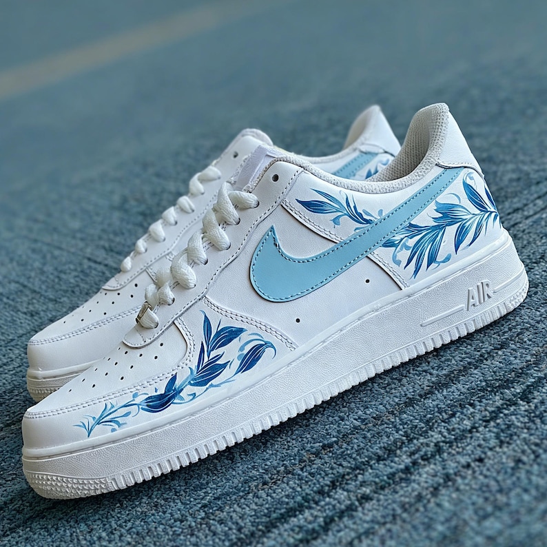 May include: White leather sneakers with light blue floral design accents. The sneakers feature a light blue Nike swoosh and matching leaf patterns. The soles are white with the word "AIR" printed on the side.