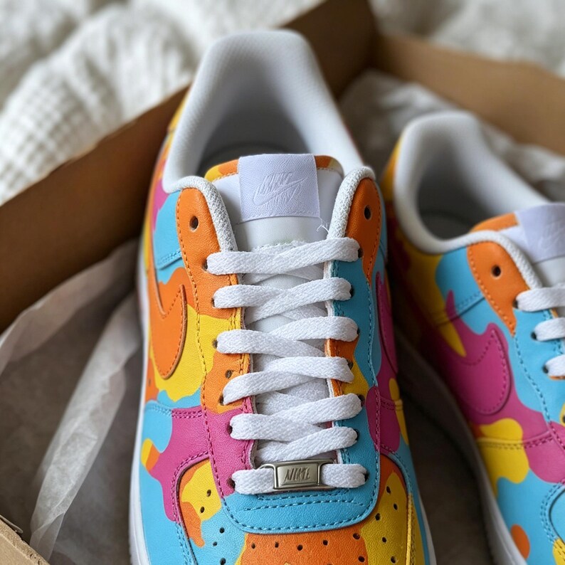 May include: A pair of colorful Nike Air Force 1 sneakers. The shoes feature a vibrant design with orange, yellow, pink, and blue camouflage-style patterns. White laces and a white tongue with the Nike logo complete the look. The shoes are in a cardboard box.