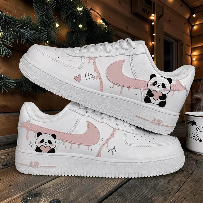 Puede incluir: Zapatillas blancas con detalles rosas, con un panda que sostiene un coraz&oacute;n y un dise&ntilde;o de pintura que gotea. Los zapatos tienen la palabra "AIR" impresa en el lateral y est&aacute;n decorados con corazones y estrellas.