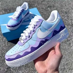 May include: Customized Air Force 1 sneakers in shades of light blue and purple with a wavy design. The shoes have white laces, a white Nike swoosh, and a white sole. One shoe is held in a hand, and another sits on a blue box.