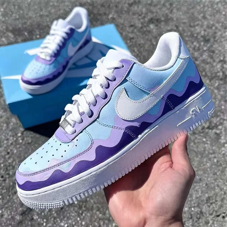 May include: Customized Air Force 1 sneakers in shades of light blue and purple with a wavy design. The shoes have white laces, a white Nike swoosh, and a white sole. One shoe is held in a hand, and another sits on a blue box.