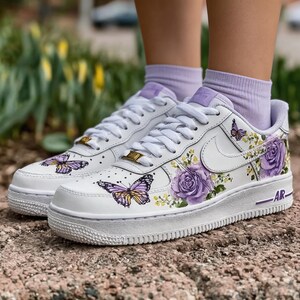 Op de afbeelding: Witte sneakers met paarse bloemen- en vlinderontwerpen. De schoenen hebben een witte zool, veters en swoosh, met gouden accenten op de tong. Het ontwerp omvat paarse rozen en gele bloemen.