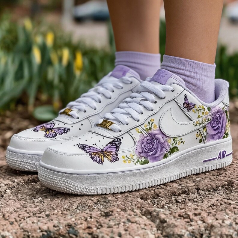 Op de afbeelding: Witte sneakers met paarse bloemen- en vlinderontwerpen. De schoenen hebben een witte zool, veters en swoosh, met gouden accenten op de tong. Het ontwerp omvat paarse rozen en gele bloemen.