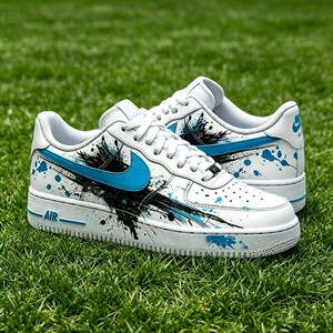 May include: White leather sneakers with blue accents and black paint splatter design. The sneakers feature a blue Nike swoosh and the word "AIR" on the side. The shoes are on a green grass background.