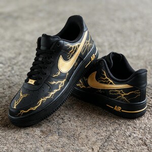 Op de afbeelding: Zwarte en gouden Air Force 1 sneakers. De schoenen hebben een zwarte basis met gouden bliksemontwerpen en gouden Nike swooshes. Het woord "AIR" is in goud op de zijkant van de zool gedrukt.