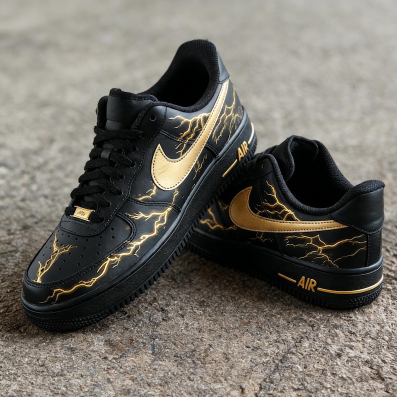 Op de afbeelding: Zwarte en gouden Air Force 1 sneakers. De schoenen hebben een zwarte basis met gouden bliksemontwerpen en gouden Nike swooshes. Het woord "AIR" is in goud op de zijkant van de zool gedrukt.