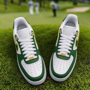 May include: Pair of white, green, and yellow athletic shoes with white laces, set on a green grassy surface. The shoes have a green trim, a yellow swoosh, and a gold accent on the laces. The background is blurred, showing a golf course.