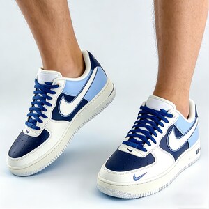 May include: Pair of white, navy blue, and light blue Air Force 1 sneakers. The sneakers feature a white Nike swoosh, navy blue laces, and a light blue heel panel. The soles are off-white.