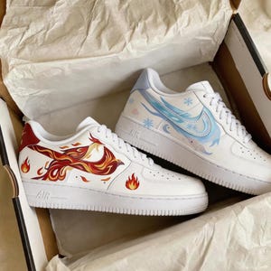 May include: White sneakers with custom artwork. One shoe features a red and orange phoenix design with flames, while the other has a blue and white winter-themed design with snowflakes and a swoosh. The shoes are in a box.