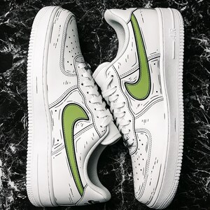 May include: Pair of white sneakers with green swoosh logos and black outline detailing. The shoes have a classic design with a white sole and laces. The sneakers are displayed on a dark, marbled surface.