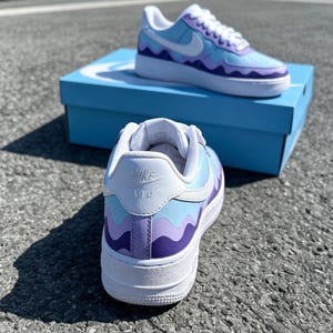 May include: Custom-painted Nike Air Force 1 sneakers in shades of blue and purple with a wavy design. The shoes have white soles and laces, and the Nike logo is visible on the side and heel. The shoes are displayed on a blue box.