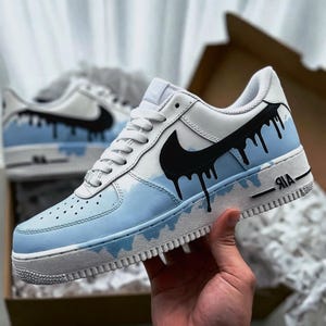 Puede incluir: Zapatillas blancas y azul claro con detalles en negro. Las zapatillas presentan un logotipo de Nike negro y un dise&ntilde;o de goteo negro &uacute;nico. La entresuela es blanca y la puntera es azul claro con peque&ntilde;as perforaciones. La palabra "AIR" est&aacute; impresa en el lateral.