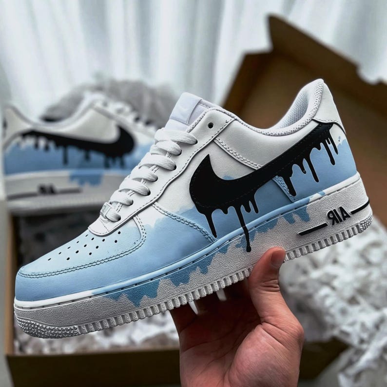 Puede incluir: Zapatillas blancas y azul claro con detalles en negro. Las zapatillas presentan un logotipo de Nike negro y un dise&ntilde;o de goteo negro &uacute;nico. La entresuela es blanca y la puntera es azul claro con peque&ntilde;as perforaciones. La palabra "AIR" est&aacute; impresa en el lateral.