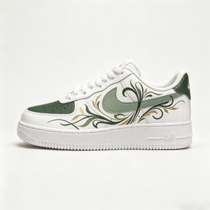 May include: White leather sneaker with green accents and a floral design. The shoe features a green Nike swoosh and a green toe cap. Gold and green floral patterns adorn the side of the shoe. The sole is white with the word "AIR" on the side.