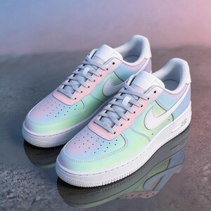May include: A pair of pastel-colored sneakers with a gradient design of pink, blue, and green hues. The shoes have white soles, laces, and a Nike swoosh. The sneakers are displayed on a reflective surface.