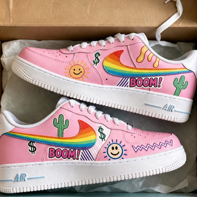 May include: Pink sneakers with white soles and laces. The shoes feature colorful graphics including a rainbow, a smiling sun, a cactus, dollar signs, and the word "BOOM!". The Nike swoosh is a rainbow design.