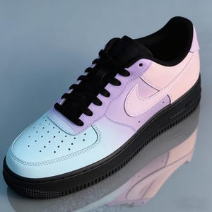 Op de afbeelding: Een enkele Air Force 1 sneaker in een verloop van pastelkleuren: lichtblauw, lavendel en roze. De schoen heeft een zwarte zool, veters en binnenvoering. De Nike swoosh is zichtbaar.