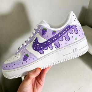 May include: White and lavender Air Force 1 sneakers with a grape-themed design. The sneakers feature a white base with lavender accents, a purple Nike swoosh, and grape illustrations. The sole is white with "AIR" printed on the side.
