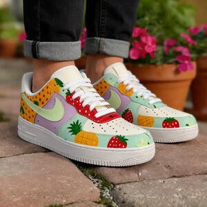 Puede incluir: Zapatillas deportivas coloridas con suela y cordones blancos. Los zapatos presentan dise&ntilde;os de frutas, como fresas y pi&ntilde;as, sobre paneles amarillos, morados y verdes. El logo de Nike es verde claro.