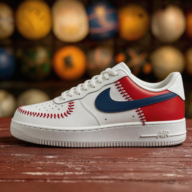 Op de afbeelding: Witte en rode honkbal-thema sneakers met blauwe accenten. De schoenen hebben rode stikseldetails, een blauwe Nike swoosh en een witte zool. Het woord "AIR" is zichtbaar aan de zijkant van de zool.
