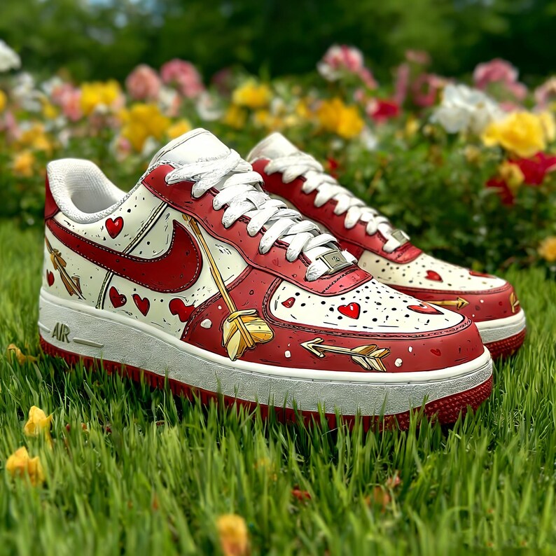 May include: A pair of red and white sneakers with heart and arrow designs. The shoes have a white base with red accents, including the Nike swoosh and a red sole. The design includes hearts and arrows, with white laces.