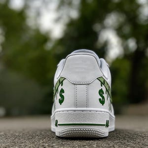 May include: White trainer with green dollar sign graphics on the heel. The shoe has a white sole and green accents. The design features a dripping dollar sign pattern.
