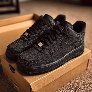May include: Pair of black Air Force 1 sneakers with a glittery, textured finish. The shoes have black laces, a black Nike swoosh, and a black sole. The sneakers are displayed on top of a brown cardboard box.