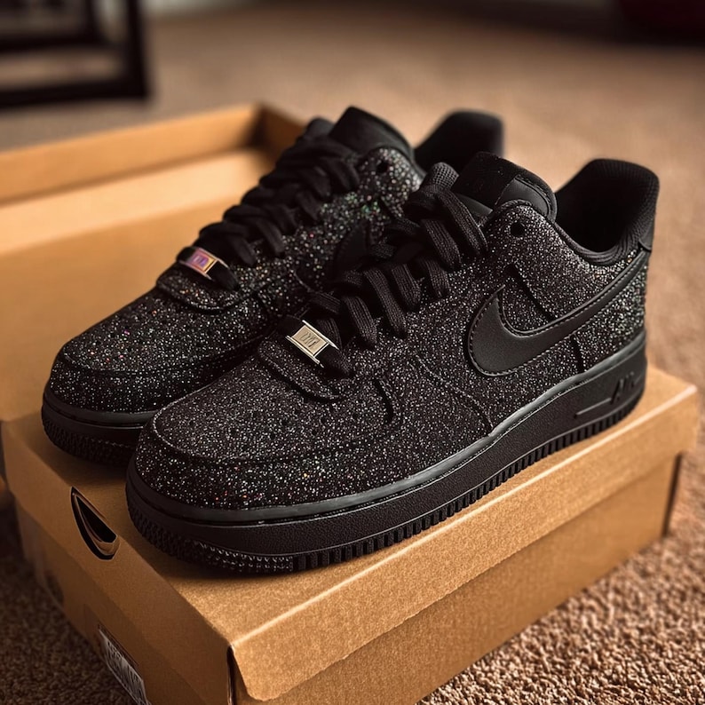 May include: Pair of black Air Force 1 sneakers with a glittery, textured finish. The shoes have black laces, a black Nike swoosh, and a black sole. The sneakers are displayed on top of a brown cardboard box.