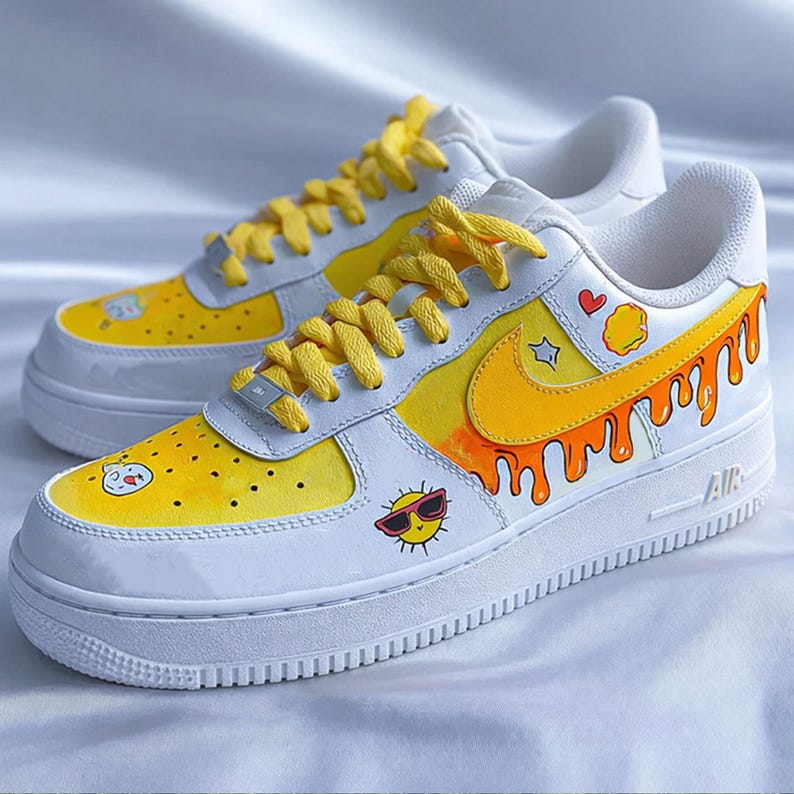 May include: A pair of white sneakers with yellow and orange accents. The shoes feature a yellow swoosh, dripping paint design, and cartoon sun graphics. Yellow laces complete the look.