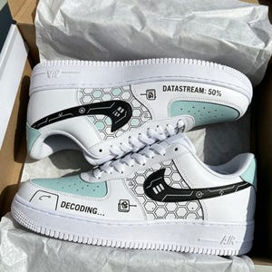 May include: White sneakers with black and light blue graphics. The shoes feature the text "DATASTREAM: 50%" and "DECODING...". The design includes a honeycomb pattern and tech-inspired illustrations. The shoes are in a white box.
