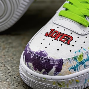 May include: White sneakers with green laces and the word "JOKER" in red lettering. The toe and sides of the shoe feature a colorful splatter paint design in purple, blue, and yellow.