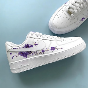 May include: White sneakers with purple floral branch designs. The shoes have a classic low-top style with white laces and soles. The word "AIR" is visible on the side of the sole. The background is a light blue.