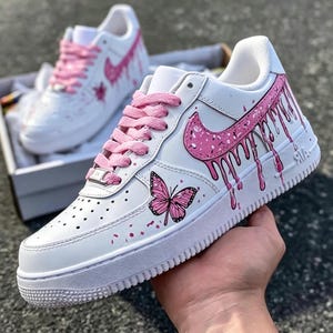 May include: White sneakers with pink accents, including a dripping design on the side and a butterfly graphic. The shoes have pink laces and a white sole. The design is repeated on the second shoe in the background.