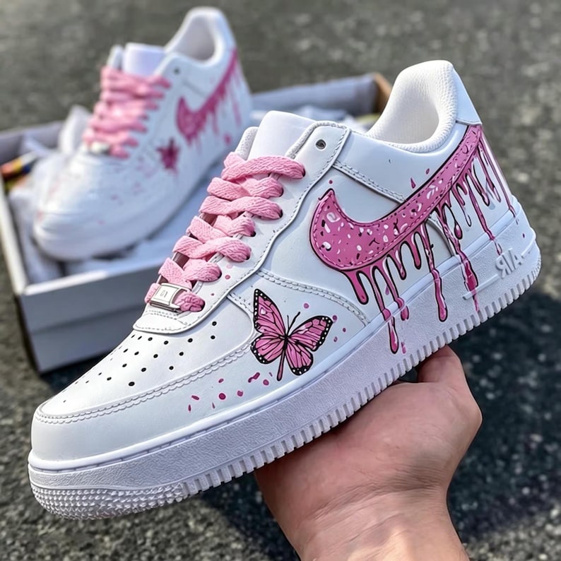 May include: White sneakers with pink accents, including a dripping design on the side and a butterfly graphic. The shoes have pink laces and a white sole. The design is repeated on the second shoe in the background.
