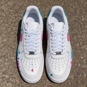 May include: Pair of white sneakers with pink and teal accents. The shoes feature a pink swoosh, teal collar, and colorful paint splatter details. White laces and a silver metal lace tag complete the design.