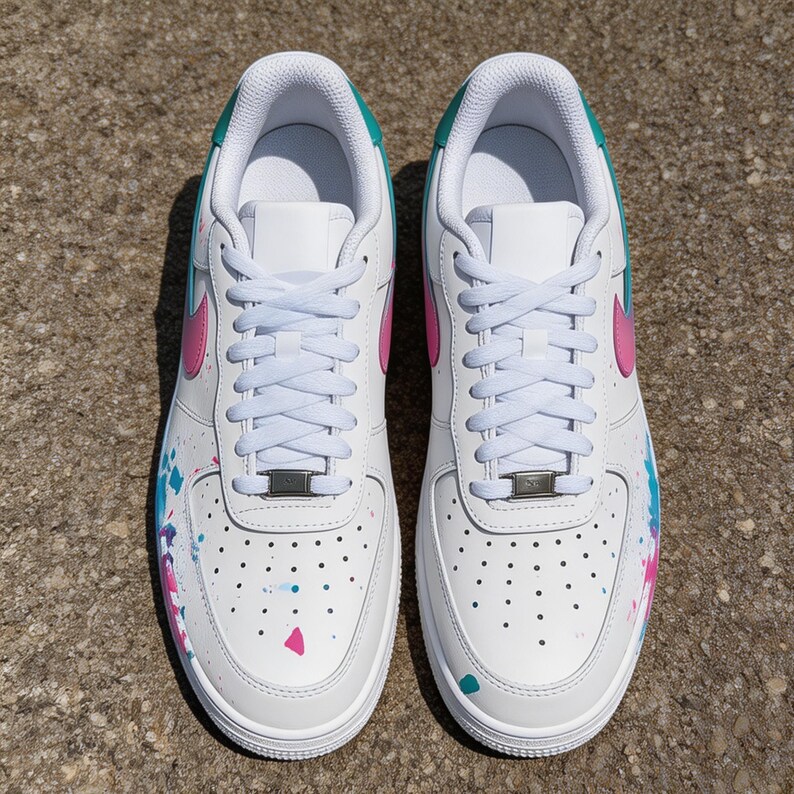 May include: Pair of white sneakers with pink and teal accents. The shoes feature a pink swoosh, teal collar, and colorful paint splatter details. White laces and a silver metal lace tag complete the design.