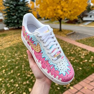 May include: A white sneaker with a custom design featuring pink and blue dripping ice cream, sprinkles, and a waffle cone. The shoe has white laces and a white sole. The design is on the toe, side, and heel of the shoe.