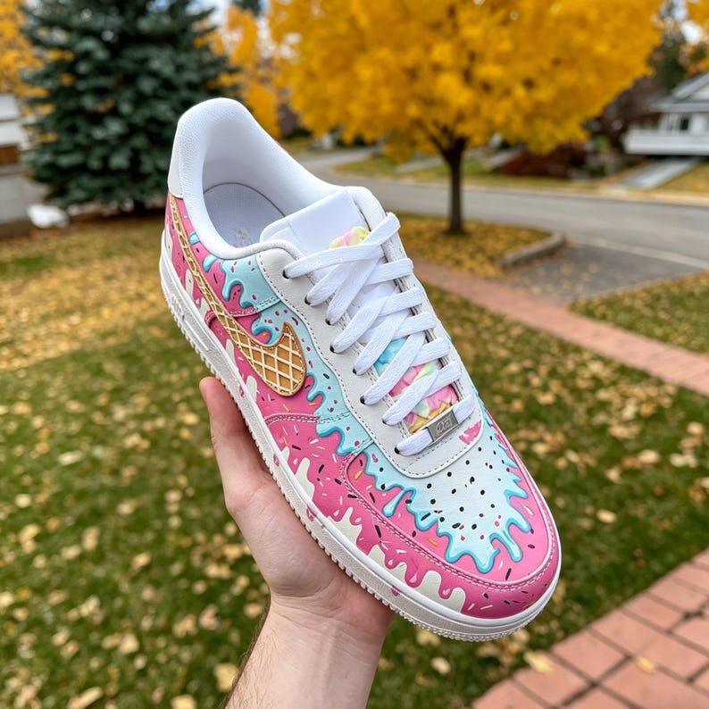 May include: A white sneaker with a custom design featuring pink and blue dripping ice cream, sprinkles, and a waffle cone. The shoe has white laces and a white sole. The design is on the toe, side, and heel of the shoe.