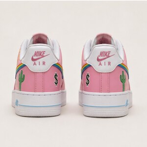 May include: Pair of pink Nike Air Force 1 sneakers. The shoes feature a white sole, a rainbow design, a green cactus, and a dollar sign. The Nike logo is visible on the back of the shoe.
