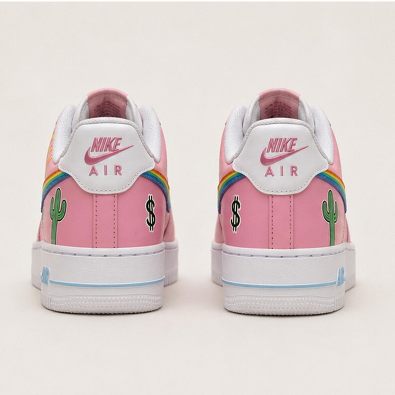 May include: Pair of pink Nike Air Force 1 sneakers. The shoes feature a white sole, a rainbow design, a green cactus, and a dollar sign. The Nike logo is visible on the back of the shoe.