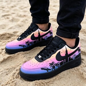 May include: Black and multi-colored sneakers with a cityscape design. The shoes feature a gradient of pink, orange, and blue hues, with a black silhouette of palm trees, buildings, and a car. The Nike swoosh is black.