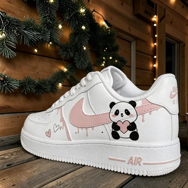 Puede incluir: Zapatilla de cuero blanco con un logotipo rosa y detalles decorativos. La zapatilla presenta un gr&aacute;fico de panda sosteniendo un coraz&oacute;n, con detalles de goteo rosa e ilustraciones de corazones. La palabra "AIR" est&aacute; impresa en rosa en la suela.