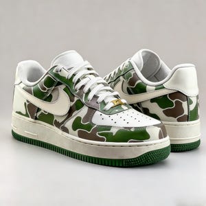 May include: Pair of white, green, and brown camouflage sneakers. The shoes feature a white leather upper with a green and brown camouflage pattern, a white swoosh, and a green sole. The laces are white, and the midsole is off-white.