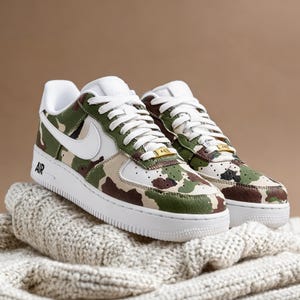 May include: A pair of camouflage-patterned sneakers with white accents, laces, and soles. The sneakers feature a green, brown, and tan camouflage design on the upper, with a white Nike swoosh and the word "AIR" on the side.