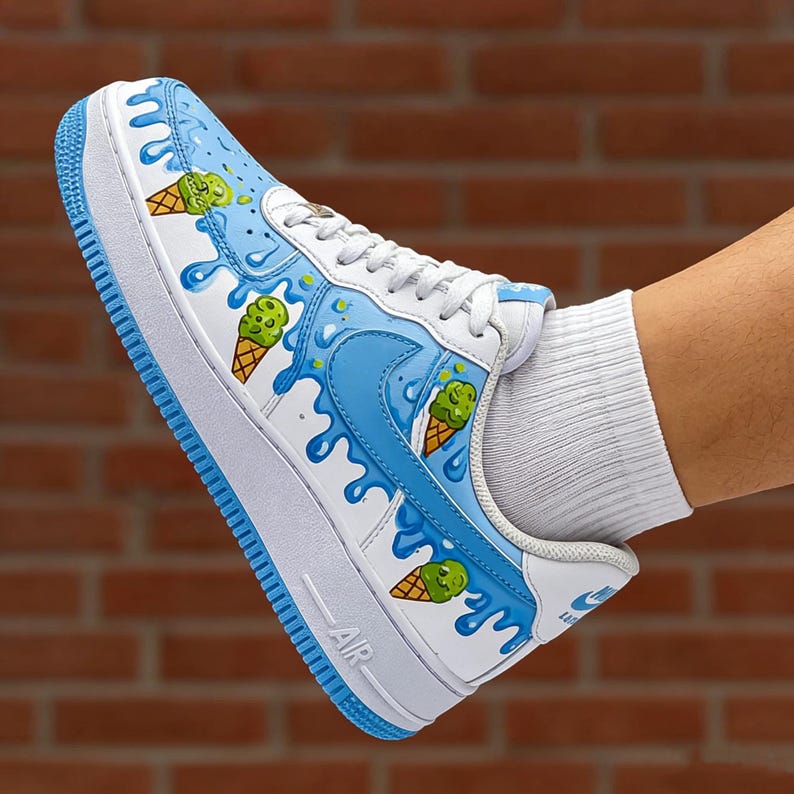 Puede incluir: Zapatillas blancas y azules con tem&aacute;tica de helado. El calzado presenta una base blanca con detalles en azul, incluyendo un dise&ntilde;o de pintura goteando y gr&aacute;ficos de conos de helado. La suela es azul y se ve la palabra "AIR".