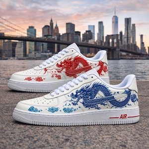 May include: A pair of white sneakers featuring a red dragon graphic on one shoe and a blue dragon graphic on the other. Both shoes have white laces, a perforated toe box, and a white sole with the word AIR printed in red on the side heel area.